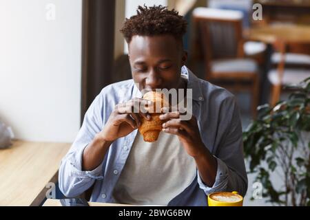 Stile di vita moderno. Uomo afro-americano che mangia croissant per pranzo in una caffetteria Foto Stock