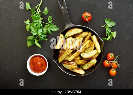 Spicchi di patate fritte con spezie in padella su fondo di pietra nera. Vista dall'alto, disposizione piatta Foto Stock