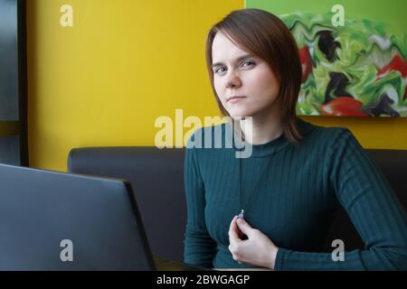 Una ragazza con un'espressione penosa di tensione sul suo viso si siede, lavorando a un computer portatile a un tavolo in un bar su un divano marrone contro una parete gialla. Guarda la macchina fotografica, tiene un gioiello sul petto con la mano. Foto Stock