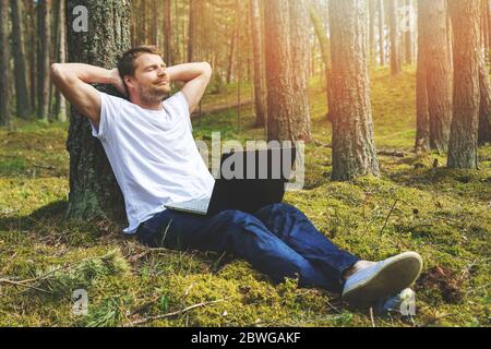 giovane uomo con computer portatile appoggiato sull'albero e rilassante nel parco Foto Stock