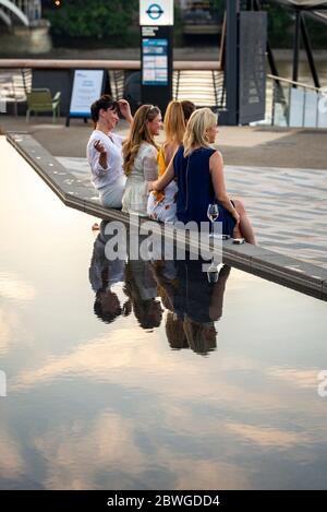 Un gruppo di donne amiche si siedono accanto a una funzione acquatica nello spazio pubblico sulla passeggiata sul lungofiume vicino alla riqualificazione della stazione di energia di Battersea, il Tamigi Foto Stock
