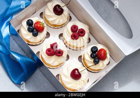 Cupcake con panna montata, ciliegie fresche, lamponi, mirtilli, fragole in scatola regalo con nastro blu. Foto Stock