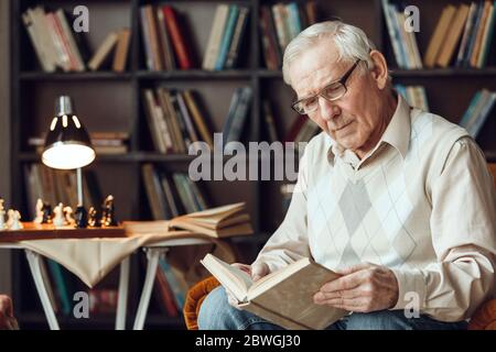Uomo anziano da solo seduto in biblioteca in occhiali lettura libro concentrato Foto Stock