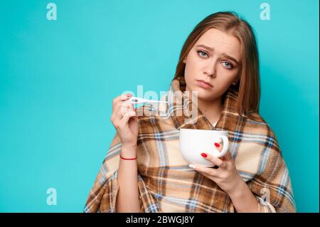 Donna in un maglione e una tazza nelle sue mani misura la temperatura con un termometro. Virus, influenza, malato, covid19, concetto di coronavirus Foto Stock