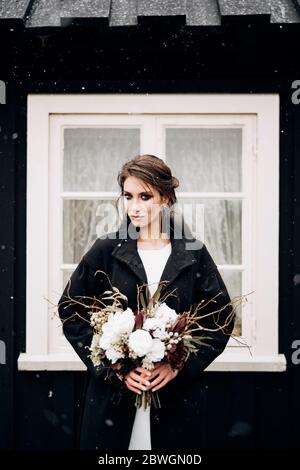 Ritratto di una sposa in un abito da sposa di seta bianca e un cappotto nero con bouquet di sposa in mano. Casa di legno nero con finestra bianca Foto Stock