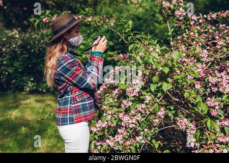 Donna fotografo scattare foto utilizzando la fotocamera in giardino estivo con maschera di coronavirus. Freelance che cammina nel parco Foto Stock