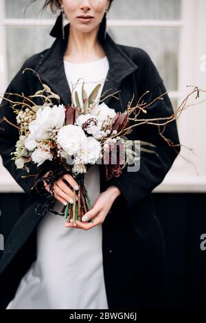 Ritratto di una sposa in un abito da sposa di seta bianca e un cappotto nero con bouquet di sposa in mano. Casa di legno nero con finestra bianca Foto Stock