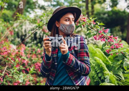 Donna fotografo scattare foto su fotocamera digitale in giardino estivo con maschera di coronavirus. Freelance ama il lavoro Foto Stock