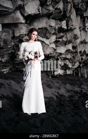 Destinazione Islanda matrimonio. La sposa in un abito di seta bianca con un bouquet in mano, si erge su sabbia nera, sulla spiaggia di Vik, sotto una roccia Foto Stock