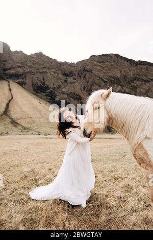 La sposa si inriga sul volto di un cavallo color crema con una lussuosa manna, in un campo di erba gialla, contro una montagna rocciosa. Destinazione Islanda Foto Stock