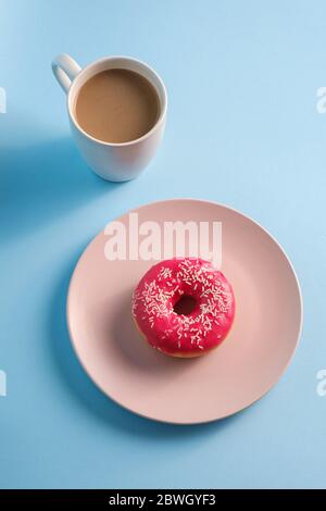 Ciambella rosa con spruzzini sul piatto vicino alla tazza di caffè, dessert con cibi dolci e bevande calde su sfondo blu minimo, vista angolare Foto Stock