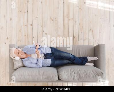 Donna anziana che ascolta la musica nelle cuffie mentre ti rilassi sul divano con lo smartphone Foto Stock