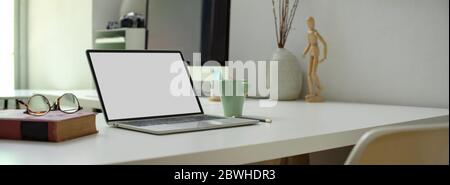 Inquadratura ritagliata di un'area di lavoro portatile con computer portatile, libro, occhiali e altri materiali di consumo per ufficio in sala ufficio Foto Stock