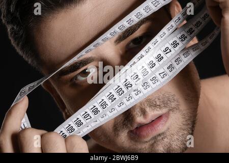 Uomo malato con nastro di misurazione su sfondo scuro, primo piano. Concetto di anoressia Foto Stock