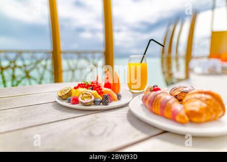 Colazione con croissant, bicchiere di succo d'arancia e frutta. Tavolo mattutino del resort tropicale con vista mare offuscata. Foto Stock