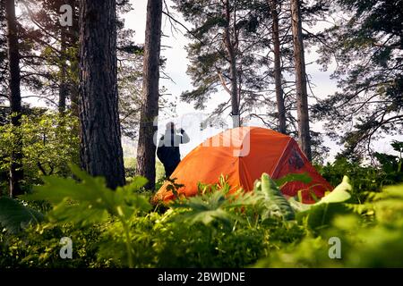 Il turista bearded sta bevendo il caffè vicino alla tenda arancione nella foresta lussureggiante alle montagne Foto Stock