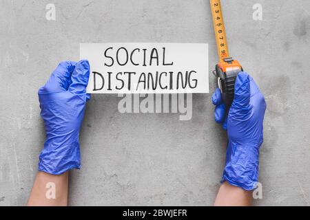 Mani in guanti di gomma che tengono carta con testo SOCIALE DISTANZIAMENTO e metro a nastro su sfondo grunge Foto Stock