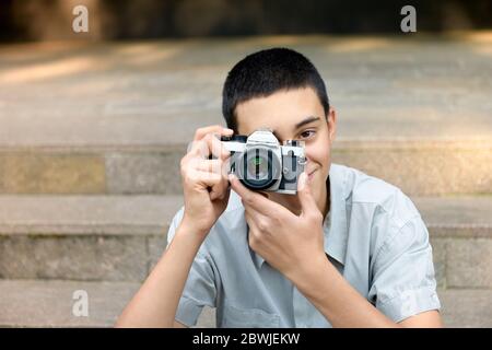 Giovane ragazzo che fotografa lo spettatore con un sorriso amichevole mentre mette a fuoco il suo scatto mentre si siede all'aperto su gradini urbani Foto Stock