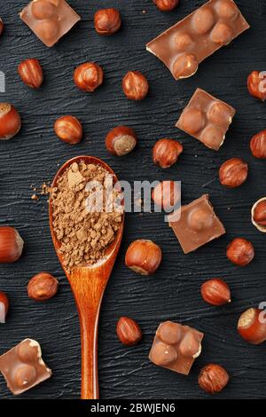 Cioccolato con nocciole e un cucchiaio di legno con cacao su fondo scuro, circondato da noci in guscio e sbucciato. Foto Stock