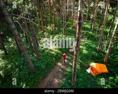 Vista aerea dell'uomo che cammina vicino alla tenda arancione nella foresta in montagna. Foto scattata con il drone Foto Stock