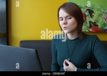 Una ragazza con un'espressione penosa di tensione sul suo viso si siede, lavorando a un computer portatile a un tavolo in un bar su un divano marrone contro una parete gialla. Tiene un gioiello sul petto con la mano. Foto Stock