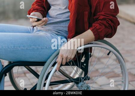 Una donna in sedia a rotelle con un telefono in mano cammina per strada, la sua mano è vicina, un irriconoscibile Foto Stock