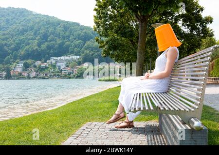 Giovane donna seduta su una panchina vicino al lago con un secchio arancione sulla testa. Sta aspettando segretamente qualcosa. Foto Stock