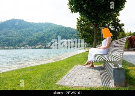 Giovane donna seduta su una panchina vicino al lago con un secchio arancione sulla testa. Sta aspettando segretamente qualcosa. Foto Stock