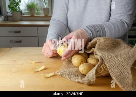 Le mani di una donna stanno peeling patate crude su un tavolo di legno in cucina a casa, fuoco selezionato, profondità di campo stretta Foto Stock