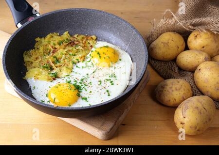 Uova fritte e roesti svizzeri in una padella su un tavolo da cucina in legno con un sacco di patate, fuoco selezionato, profondità di campo stretta Foto Stock