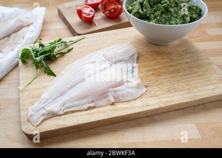 Filetto di passera cruda, pomodori e una ciotola di sedano e rucola schiacciati su un tavolo da cucina in legno, preparazione per cucinare panini di pesce, selezionati f Foto Stock