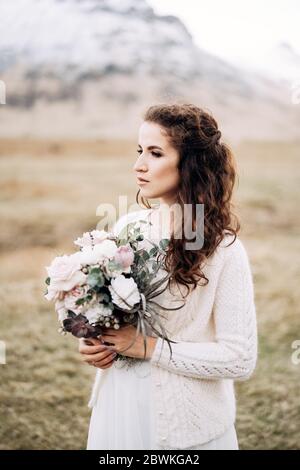 Ritratto di una sposa in un abito da sposa bianco, con bouquet di sposa in mano. In un campo di erba secca gialla, in mezzo a una montagna nevosa e pascolo Foto Stock