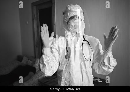Immagine in bianco e nero del medico in tuta PPE, respiratore, guanti, con fonendoscopio, paziente in visita a casa durante la quarantena covid-19, dopo aver disinfettato le mani per essere pronti per l'esame Foto Stock
