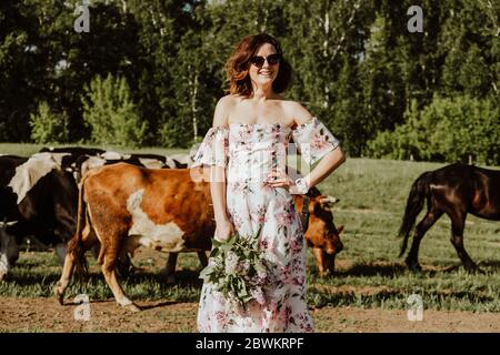 Ritratto esterno atmosfera stile di vita foto di giovane bella donna dai capelli scuri in un abito rosa si erge sullo sfondo di un gregge di mucche. Ragazza Foto Stock