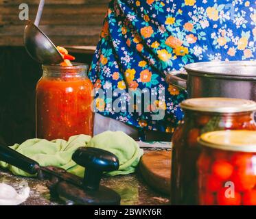 Un uomo in un grembiule colorato zoccoli pomodori e Lecho salsa in vasetti di vetro in un casale Foto Stock