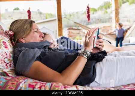 Madre e figlia si stendono insieme su un letto esterno che guarda uno schermo del telefono intelligente Foto Stock