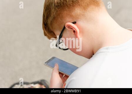 Vista da dietro di un ragazzo dai capelli rossi con cellulare e occhiali in un contesto urbano. Gli occhiali da ragazzo consentono di tenere lo smartphone a portata di mano. Foto Stock