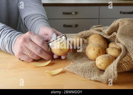 Le mani di una donna stanno peeling patate crude con un vecchio pelatore su un tavolo di cucina in legno, fuoco selezionato, profondità stretta di campo Foto Stock