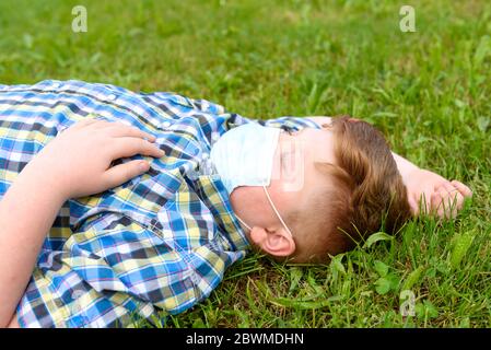 Ritratto di un ragazzo con maschera chirurgica adagiata sull'erba del parco. Primo piano di un ragazzino in una camicia a plaid che si rilassa sull'erba. Foto Stock
