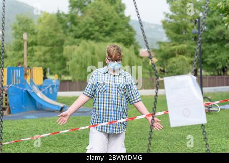 Ragazzo che indossa occhiali di protezione chirurgici vestiti in una camicia di plaid davanti ad un nastro di barriera a strisce rosse e bianche intorno al parco giochi. Foto Stock