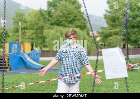 Ragazzo che indossa occhiali di protezione chirurgici vestiti in una camicia di plaid davanti ad un nastro di barriera a strisce rosse e bianche intorno al parco giochi. Foto Stock