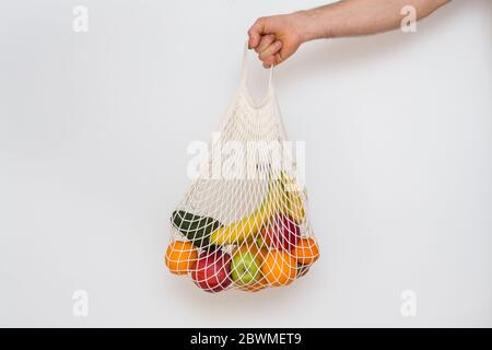 Concetto di zero sprechi con spazio di copia. Mano da uomo che tiene una borsa della spesa in rete con prodotti. Acquirente eco-compatibile in mesh. Sfondo bianco, primo piano. Plas Foto Stock