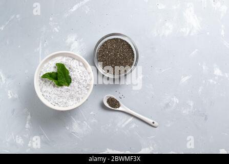 Budino di Chia con foglie di menta in una ciotola bianca e una ciotola di vetro con grani su sfondo grigio. Vista dall'alto. Una sana colazione consiste di yogurt e. Foto Stock