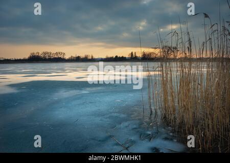 Un lago scongelamento con canne e nuvole scure dopo il tramonto sul cielo, vista invernale serale Foto Stock