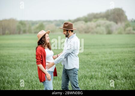 Ritratto di una donna incinta con il suo ragazzo vestito casualmente con cappelli in piedi insieme sul campo verde. Coppia felice che si aspetta un bambino, giovane concetto di famiglia Foto Stock
