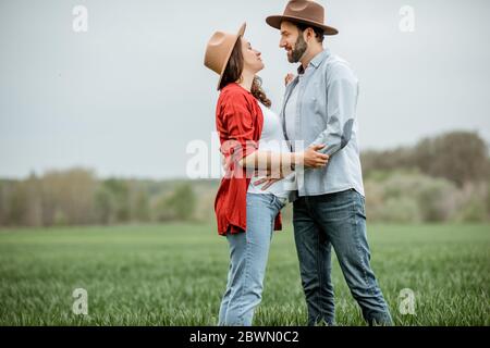 Ritratto di una donna incinta con il suo ragazzo vestito casualmente con cappelli in piedi insieme sul campo verde. Coppia felice che si aspetta un bambino, giovane concetto di famiglia Foto Stock