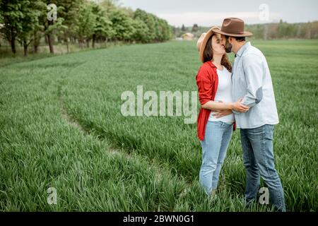 Ritratto di una donna incinta con il suo ragazzo vestito casualmente con cappelli in piedi insieme sul campo verde. Coppia felice che si aspetta un bambino, giovane concetto di famiglia Foto Stock