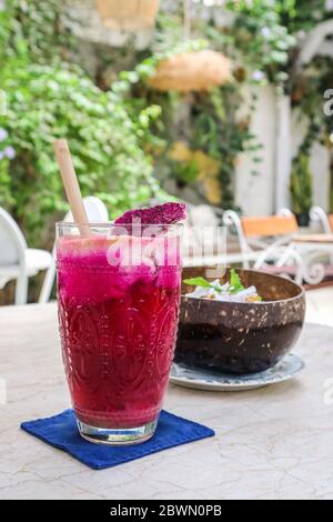 Succo rosa con mele, barbabietole, frutta drago e lime in vetro con cannuccia di bambù e recipiente per frullati sul tavolo al caffè Foto Stock