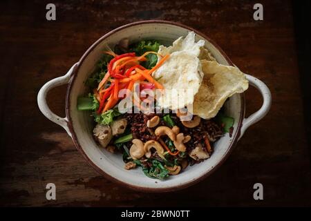 Riso Rosso biologico Nasi Goreng, riso vegetariano fritto con un sacco di verdure, tofu e anacardi tostati in ciotola su sfondo di legno, vista dall'alto Foto Stock