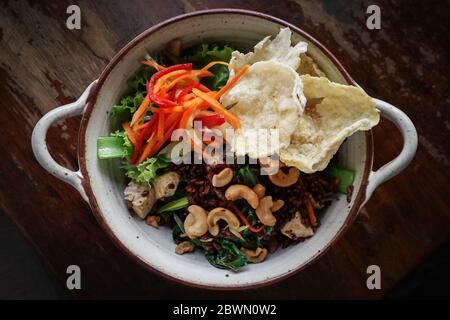 Riso Rosso biologico Nasi Goreng, riso vegetariano fritto con un sacco di verdure, tofu e anacardi tostati in ciotola su sfondo di legno, vista dall'alto Foto Stock